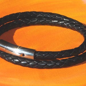 May include: Black braided leather bracelet with a silver-tone clasp. The bracelet is wrapped twice, creating a layered effect. The background is a vibrant orange.