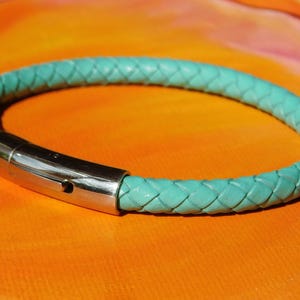 May include: A turquoise braided leather bracelet with a silver-colored magnetic clasp. The bracelet is circular and rests on an orange surface. The braided design is visible throughout the bracelet.