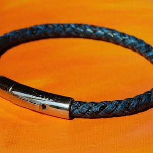 May include: A braided blue leather bracelet with a silver clasp. The bracelet is circular and set against an orange background. The clasp is rectangular and has a small hole.
