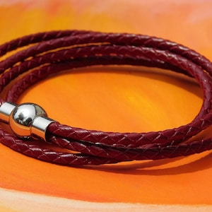 May include: A burgundy leather wrap bracelet with a braided design. The bracelet wraps multiple times and is secured with a silver magnetic clasp. The bracelet is displayed on an orange surface.