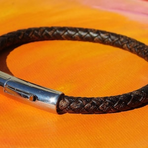 May include: Braided brown leather bracelet with a silver clasp. The bracelet is circular and lies on an orange surface. The clasp is a shiny, metallic silver. The leather is a dark brown color.