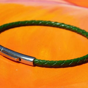 Könnte beinhalten: Ein grünes geflochtenes Lederarmband mit einem silberfarbenen Verschluss. Das Armband ist kreisförmig und liegt auf einer orangefarbenen Oberfläche. Das geflochtene Design ist sichtbar.