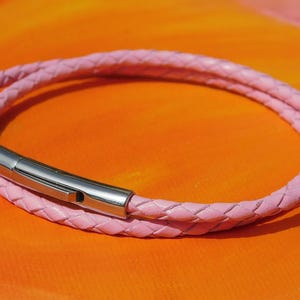 May include: A pink braided leather bracelet with a silver-colored clasp. The bracelet is wrapped around itself, creating a double-strand effect. The background is a gradient of orange and pink.
