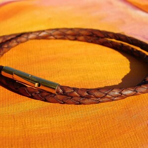 May include: A brown braided leather bracelet with a gold-colored clasp. The bracelet is wrapped around itself, creating a double-strand effect. The leather has a natural, textured appearance.