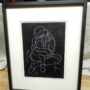 May include: Framed black and white artwork depicting a seated figure in a contemplative pose. The artwork features a minimalist style with white lines against a black background. The piece is framed in a black frame with a white mat.