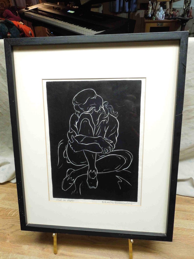 May include: Framed black and white artwork depicting a seated figure in a contemplative pose. The artwork features a minimalist style with white lines against a black background. The piece is framed in a black frame with a white mat.