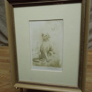 May include: A framed etching print featuring a cat looking upward at a small butterfly. The artwork is presented in a light-colored mat with a textured gold-toned frame. The print includes a signature at the bottom right corner of the image area.