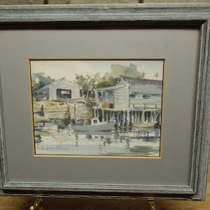 May include: Watercolor painting of a coastal scene with buildings on stilts and a small boat in the water. The artwork is framed in a gray wooden frame with a gray mat. The artist's signature is visible in the bottom left corner.