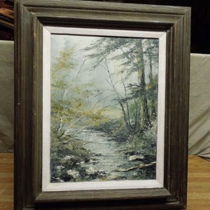 May include: Oil painting of a forest scene with a stream. The artwork is framed in a dark brown wooden frame with a white inner border. The painting depicts trees, water, and a misty atmosphere, with muted greens and browns.