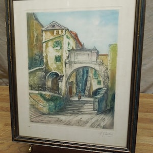 May include: Framed watercolor painting of a European street scene. The artwork depicts buildings with arched doorways and a person walking down steps. The frame is dark with gold accents, displayed on a gold easel.