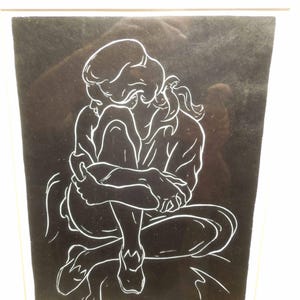May include: A framed black and white line drawing of a person in a seated position, hugging their knees. The artwork features a minimalist style with white lines against a black background, creating a striking contrast. The person's hair is pulled back in a ponytail.