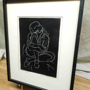 May include: Black and white artwork of a person in a seated, curled position. The artwork is in a black frame with a white mat. The frame is supported by a gold-colored stand. The artwork is signed by the artist.