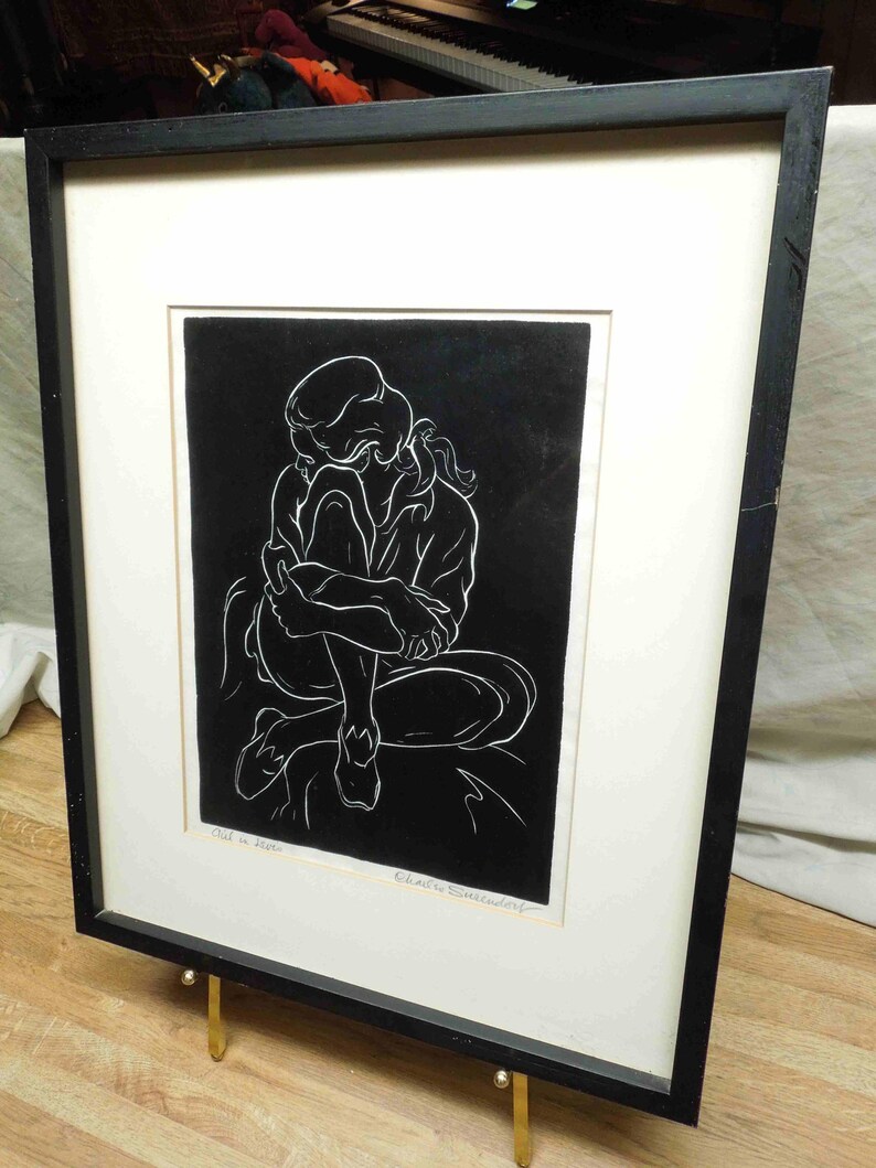 May include: Black and white artwork of a person in a seated, curled position. The artwork is in a black frame with a white mat. The frame is supported by a gold-colored stand. The artwork is signed by the artist.