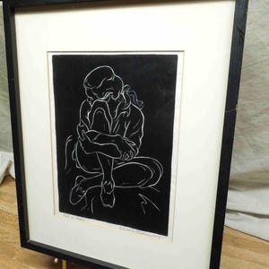 May include: A framed black and white artwork depicting a seated figure in a contemplative pose. The artwork is framed in a black frame with a white mat. The artwork is displayed on a gold easel.