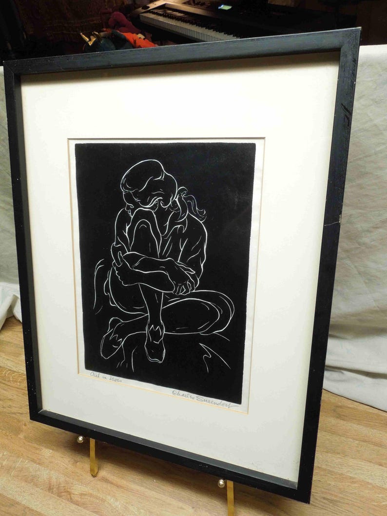 May include: A framed black and white artwork depicting a seated figure in a contemplative pose. The artwork is framed in a black frame with a white mat. The artwork is displayed on a gold easel.