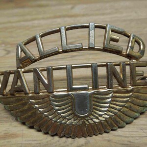 May include: Vintage Allied Van Lines emblem, featuring a gold-toned metal design. The emblem has the words "ALLIED" arched above "VAN LINES", with a stylized wing design below. The emblem is set against a wooden surface.