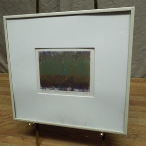 May include: A framed abstract art print with a white border. The artwork features a rectangular composition with green and brown hues. The frame is supported by a gold-colored easel, set against a wooden surface.