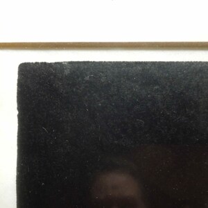 May include: Close-up of a framed artwork. The artwork features a textured black surface, possibly velvet or a similar material, set within a white mat and a light brown wooden frame. The composition is simple and abstract.