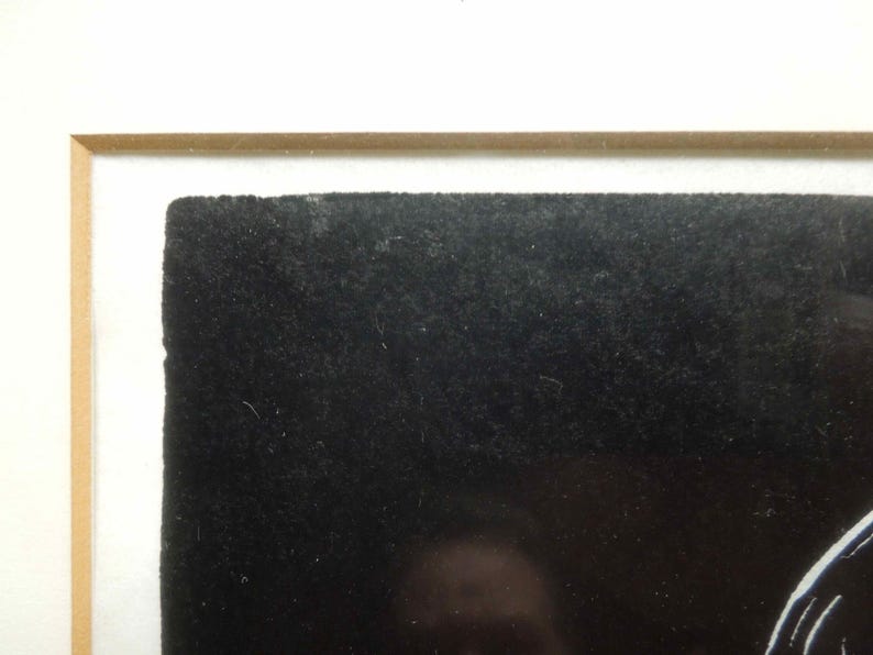May include: Close-up of a framed artwork. The artwork features a textured black surface, possibly velvet or a similar material, set within a white mat and a light brown wooden frame. The composition is simple and abstract.