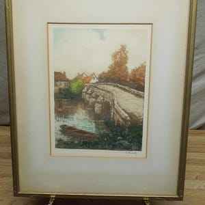 May include: Framed landscape art print featuring a stone bridge over a river, with a small boat and houses in the background. The artwork is in a gold-colored frame and displayed on a brass easel.