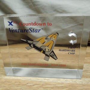 May include: Clear acrylic block with a VentureStar spacecraft design in yellow, gray, and black. Text includes "Countdown to VentureStar," "2nd Anniversary - July 1998," and "First Hardware Chip."