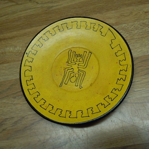 May include: A yellow ceramic plate with a black spider design in the center. The plate has a black border with a repeating pattern.
