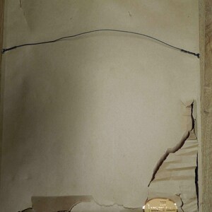 May include: The back of a framed artwork, showing a tan paper backing with a wire for hanging. The paper is torn in several places, revealing the frame's structure. A gold label with text is affixed to the lower right corner.