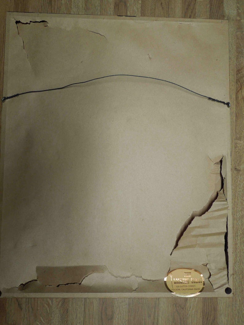 May include: The back of a framed artwork, showing a tan paper backing with a wire for hanging. The paper is torn in several places, revealing the frame's structure. A gold label with text is affixed to the lower right corner.