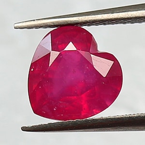 May include: A heart-shaped, faceted ruby gemstone held by tweezers. The ruby is a deep, vibrant red colour with multiple facets that catch the light. The gemstone is set against a blurred, neutral background.