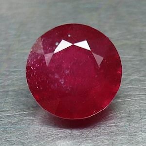 May include: A round, red gemstone with a faceted cut. The gemstone is set against a gray background.