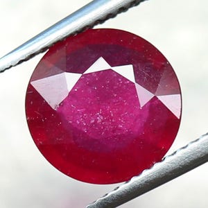Könnte beinhalten: Ein runder, facettierter Rubin-Edelstein in tiefroter Farbe. Der Rubin wird in einer Pinzette gehalten.