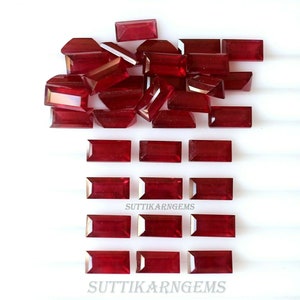 May include: A pile of loose red ruby gemstones and a row of 10 rectangular cut red ruby gemstones. The gemstones are arranged on a white surface. The text "SUTTIKARN GEMS" appears twice on the surface.