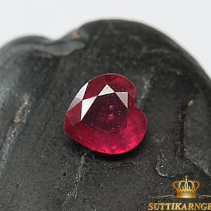 Könnte beinhalten: Ein herzförmiger, facettierter Rubin-Edelstein in tiefroter Farbe. Der Edelstein wird auf einer dunklen, strukturierten Oberfläche präsentiert. Das Bild enthält den Text "SUTTIKARNGEMS gems for jewelry" in Gold.
