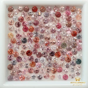 Puede incluir: Una colección de piedras preciosas sueltas de color rosa, rojo y blanco en una caja cuadrada blanca. Las piedras preciosas son redondas y tienen una variedad de tamaños. La caja está etiquetada con "SUTTHIARN GEMS".