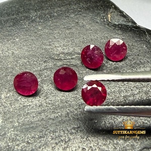 Pode incluir: Cinco rubis redondos, facetados e de cor vermelho profundo. Um rubi é segurado por uma pinça de metal prateado, enquanto os outros quatro estão dispostos por perto. O texto SUTTIKARNGEMS gems for jewelry é visível no canto inferior direito.