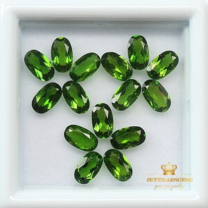 May include: A collection of 12 oval-shaped green gemstones, likely peridot, displayed in a white square box. The gemstones are arranged in a flower-like pattern. The text "SUTTIKARN GEMS gems for jewelry" is visible in the bottom right corner of the image.
