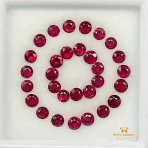 May include: A collection of twenty-four round, faceted, deep red rubies arranged in two concentric circles within a white display case. The bottom right corner features a gold crown icon above the text SUTTIEARNGEMS gems for jewellery.