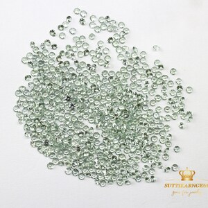 May include: A large quantity of small, round, light green gemstones. The gemstones are scattered on a white surface.