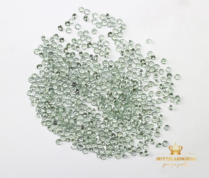 May include: A large quantity of small, round, light green gemstones. The gemstones are scattered on a white surface.