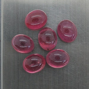Pode incluir: Seis pedras preciosas ovais de cor rosa com um leve brilho. As pedras preciosas estão dispostas em semicírculo sobre uma superfície prateada.