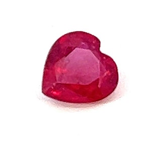 May include: A heart-shaped, faceted ruby gemstone. The ruby is a deep, saturated red color, with a polished surface that reflects light. The gemstone is set against a white background.
