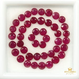 May include: A collection of round, faceted, deep red rubies arranged in a circular pattern within a white square display tray. The stones exhibit a consistent brilliant cut and vibrant colour. The text SUTTIEARNGEMS is printed in the bottom corner.