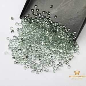 May include: A large pile of light green gemstones, scattered on a white surface. The gemstones are round and have a smooth, polished finish. The gemstones are spilling out of a black tray.