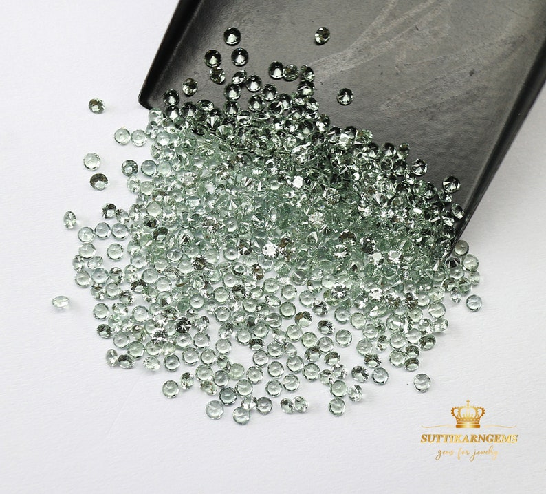 May include: A large pile of light green gemstones, scattered on a white surface. The gemstones are round and have a smooth, polished finish. The gemstones are spilling out of a black tray.