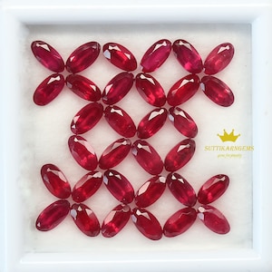 Può includere: Una collezione di 25 pietre preziose rosse di forma ovale e sfaccettate. Le pietre preziose sono disposte in una griglia in una scatola bianca. La scatola ha uno sfondo bianco e un bordo bianco. Il testo "SUTTIKARNGEMS gems for jewelry" è stampato nell'angolo in basso a destra della scatola.