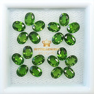 May include: A collection of 14 oval-shaped green gemstones, likely peridot, displayed in a white box. The gemstones are arranged in a symmetrical pattern, with some overlapping. The box has a white background and a white border.