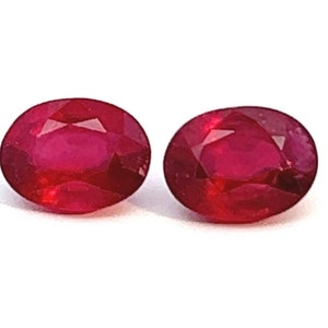 May include: Two oval-shaped red gemstones, likely rubies, are shown on a white background.