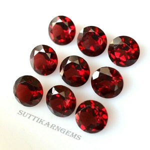 Può includere: Otto pietre preziose rotonde di colore rosso, probabilmente granati, sono disposte su una superficie bianca. Le pietre preziose sono sfaccettate e hanno una finitura lucida e lucidata. Il testo "SUTTIKARN GEMS" è visibile nell'angolo in basso a sinistra dell'immagine.