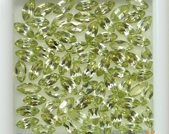 May include: A collection of light green, marquise-cut gemstones. The gems are arranged closely together within a white square, creating a dense, sparkling display. The stones have a translucent quality, reflecting light.
