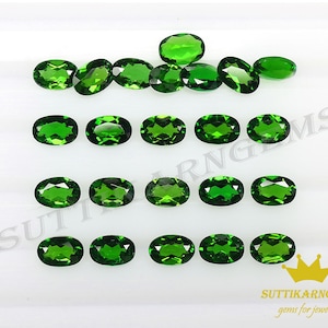 May include: Twenty-one oval-shaped green gemstones, likely emeralds, arranged in three rows on a white surface. The gemstones are all approximately the same size and shape. The image is taken from a slightly overhead angle.